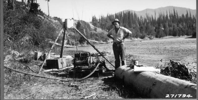 man standing with pump machinery next to a river with trees and mountains in the distance