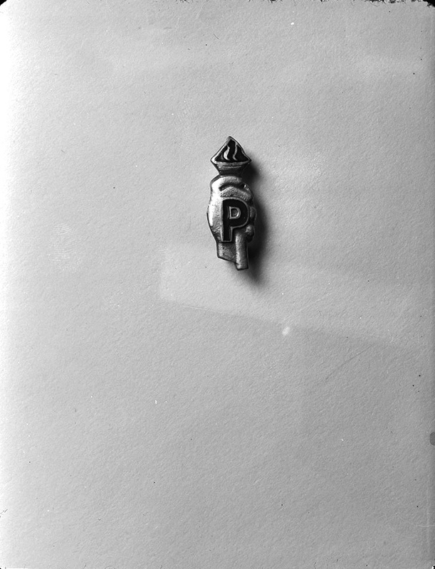 Negative photograph of a pin worn by members of Psychiana. Pin includes a small hand holding a lit torch with the capital letter 'P' prominently in front.