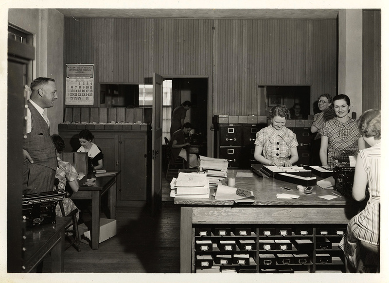 Photograph depicts young Frank B. Robinson standing in a doorway with his hands on his hips and watching smiling female Psychiana staff complete tasks.